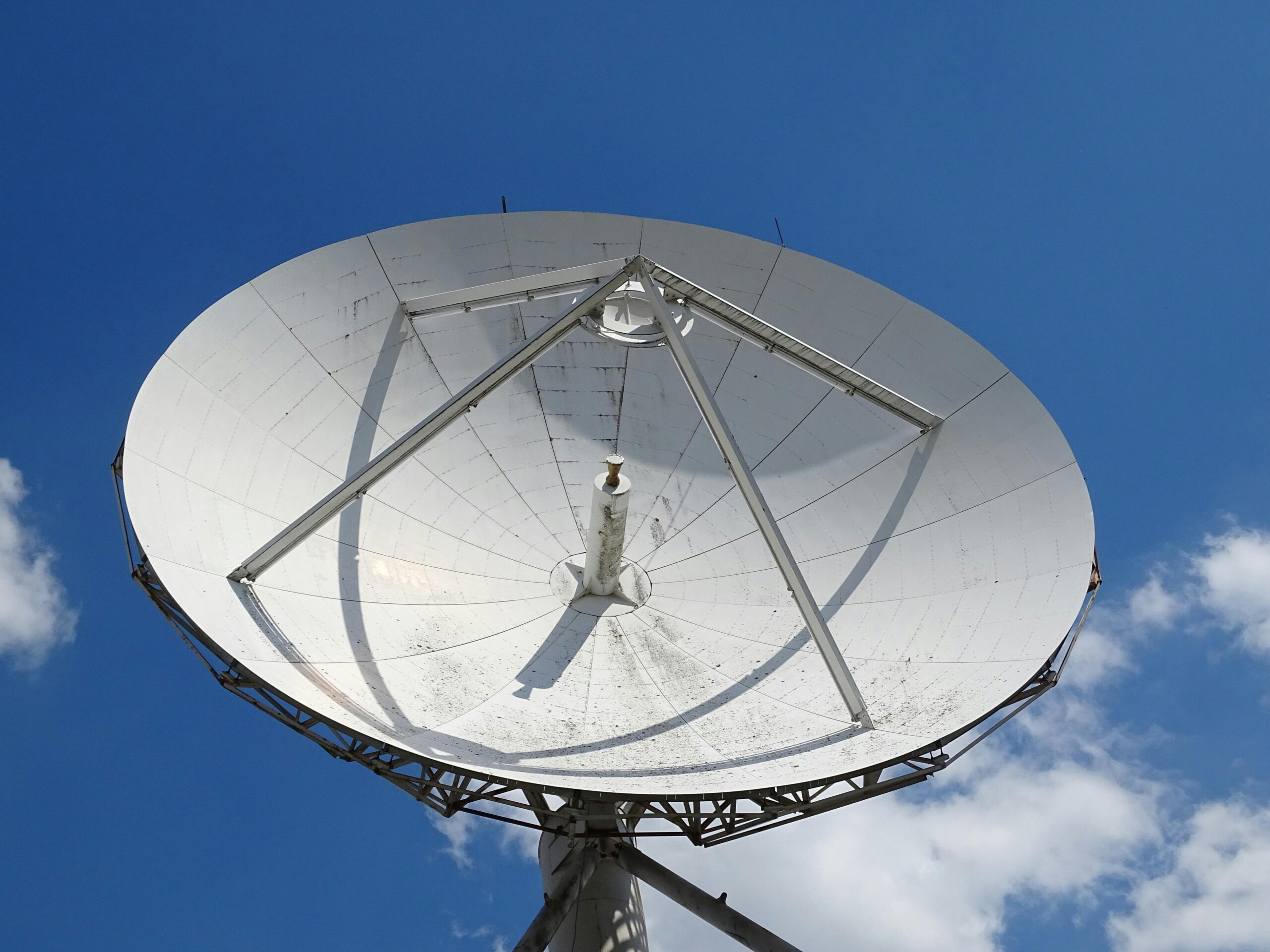 A low angle view of a large satellite dish against a clear blue sky with scattered clouds.